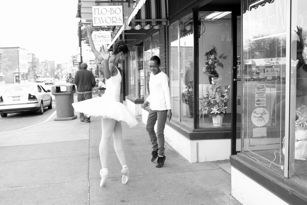 Aesha Ash in a white tutu and pointe shoes on city block. A woman dances next to her high on the tips of her sneakers.